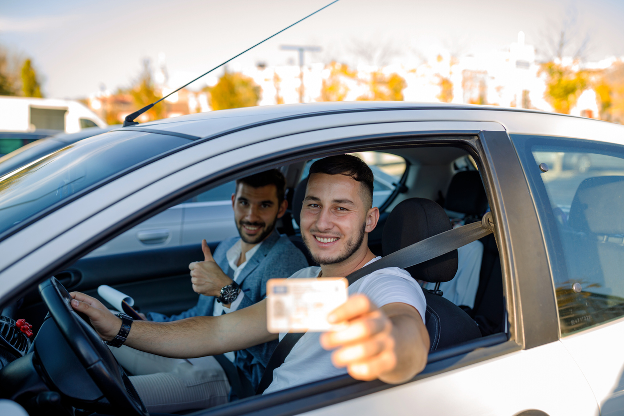 A young Caucasian male driving instructor holding thumbs-up and a young Caucasian man holding a driver's licence are sitting in a car and looking at the camera with smiles on their faces.