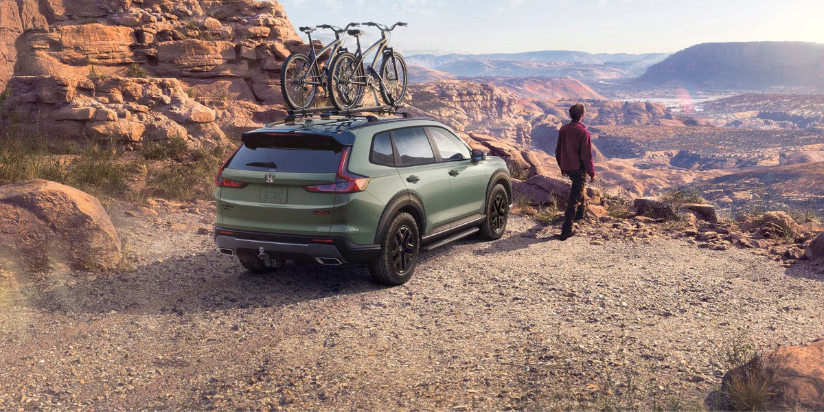 Green 2027 Honda CR-V with two bikes on a roof rack, parked on a gravel overlook as the driver takes in a sweeping canyon view
